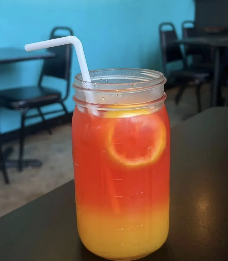 A mason jar of red and orange Kool-Aid with a lemon slice and a straw, on a table inside Wally&rsquo;s.