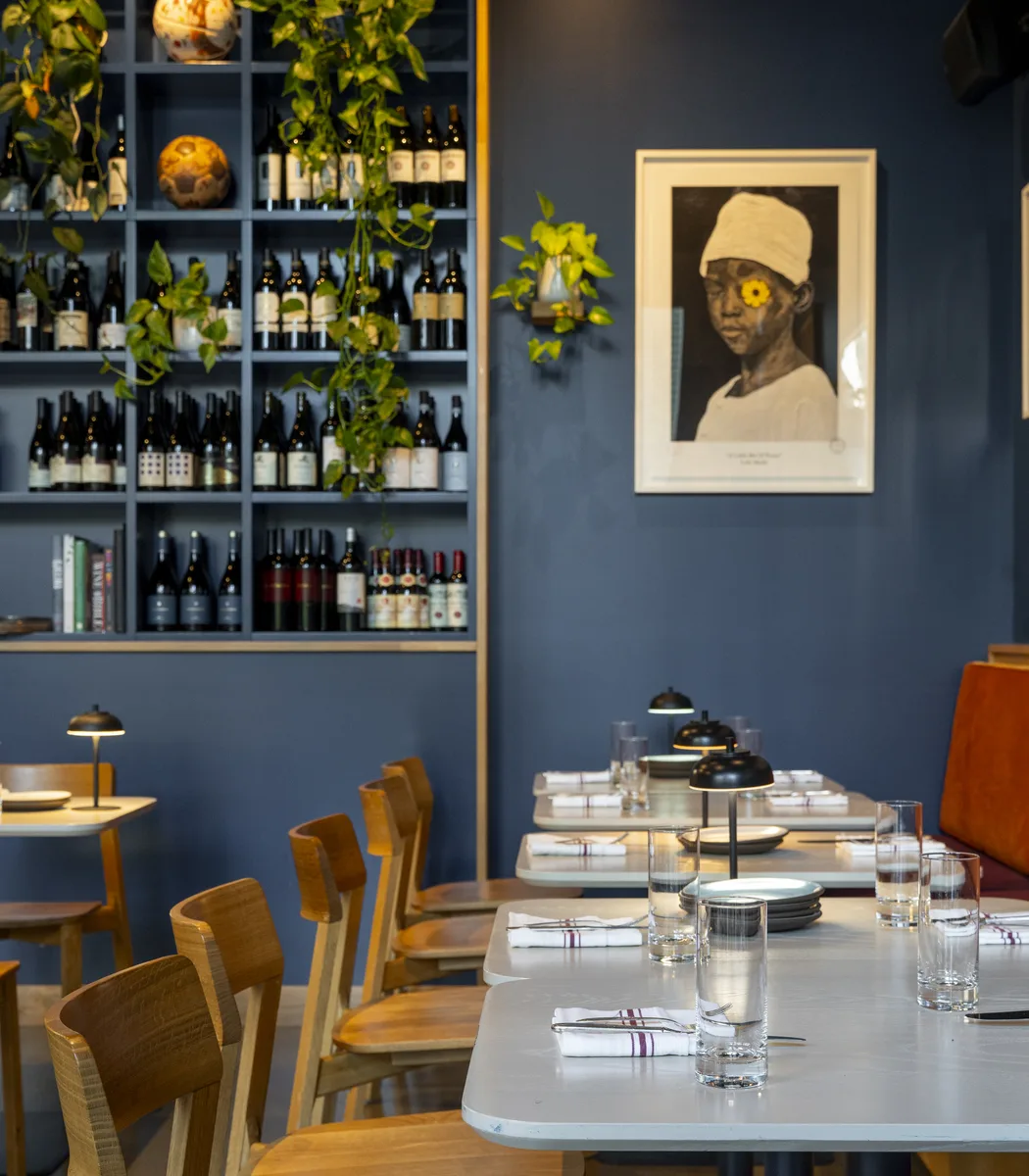 Marjorie's dining room: deep blue walls, wine shelves draped with trailing plants, wooden chairs at white tables, and a framed portrait print of a child with a yellow flower over one eye.