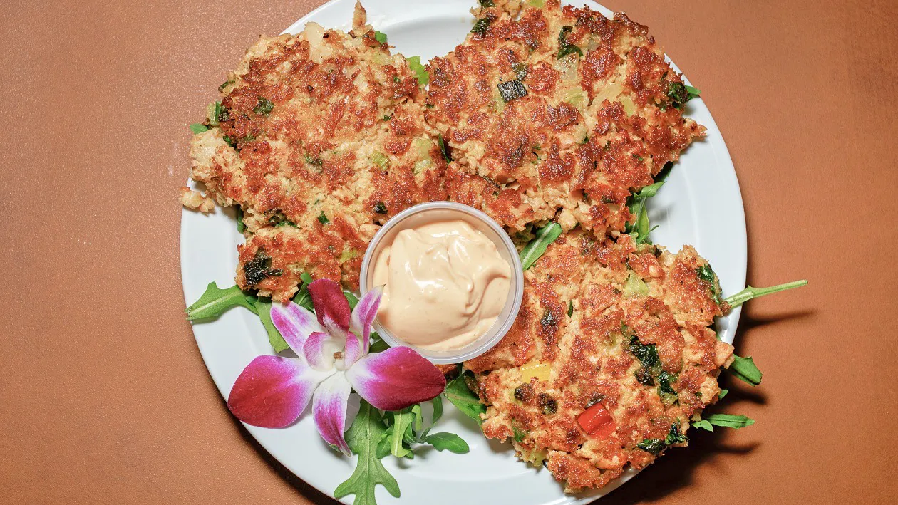 Three pan-seared salmon cakes on arugula with a cup of spicy mayo and an orchid, seen from above.