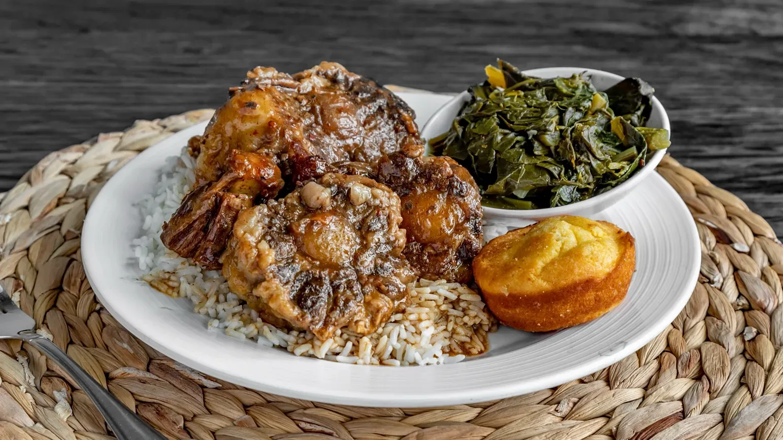 Braised oxtails in gravy over white rice, with a bowl of collard greens and a cornbread muffin, on a woven placemat.