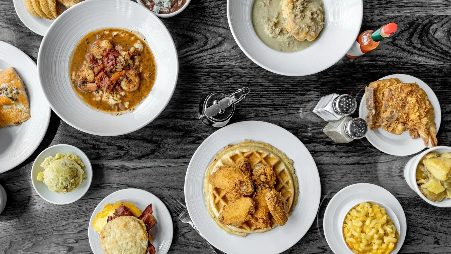 An overhead spread of soul food on a grey wood table &mdash; fried chicken and waffles, shrimp and grits, biscuits and gravy, fried catfish, red beans and rice, mac and cheese and candied yams.