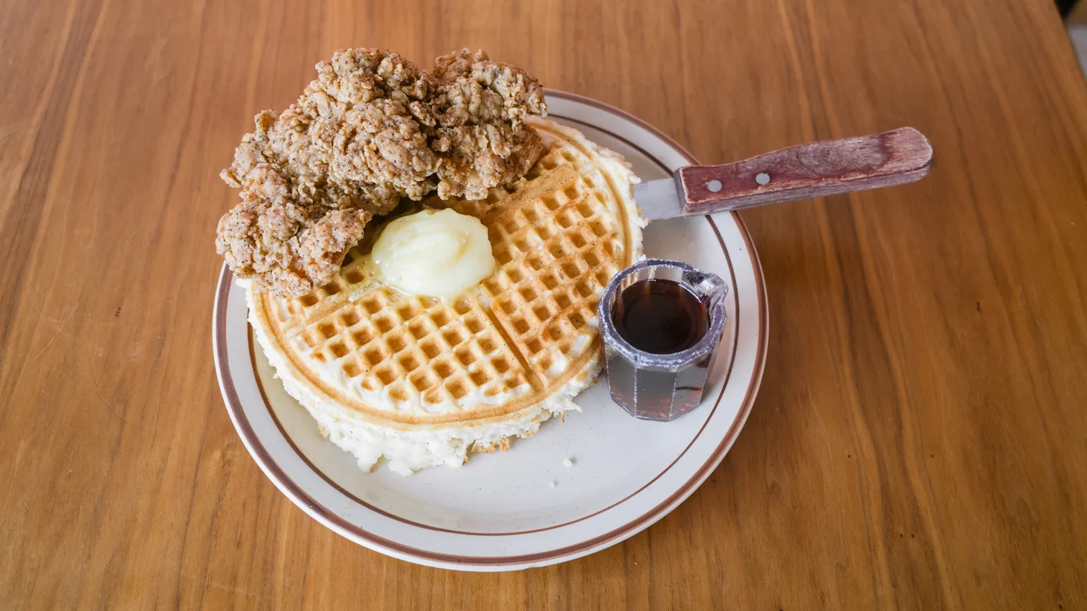 Fried chicken on a waffle with a scoop of butter and a small pitcher of syrup.