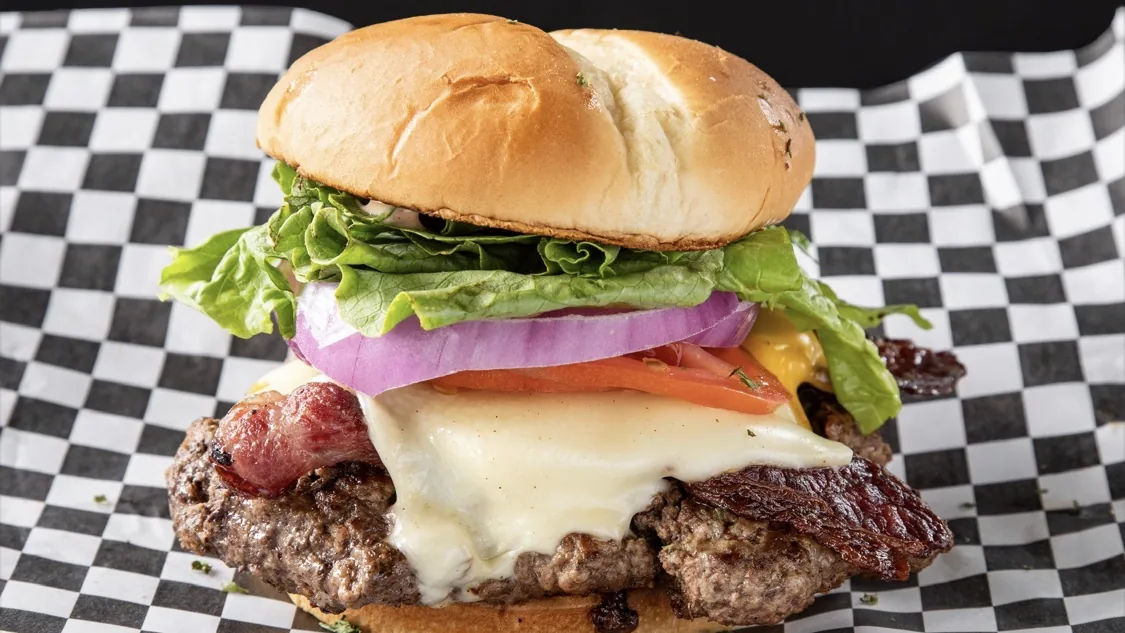 A bacon cheeseburger with melted white cheese, tomato, red onion and lettuce on a toasted bun, set on checkered paper.