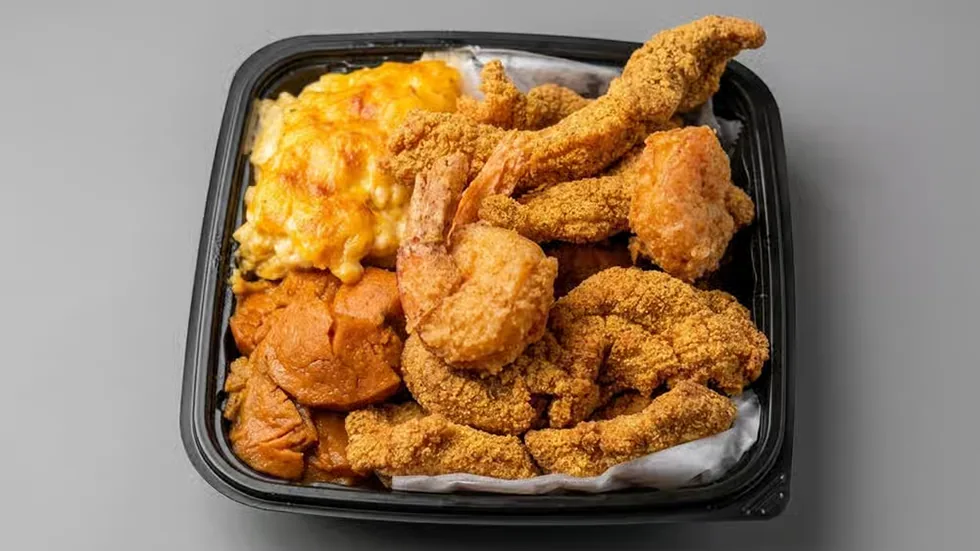 A takeout tray of crispy fried fish and shrimp with baked macaroni and cheese and candied yams.