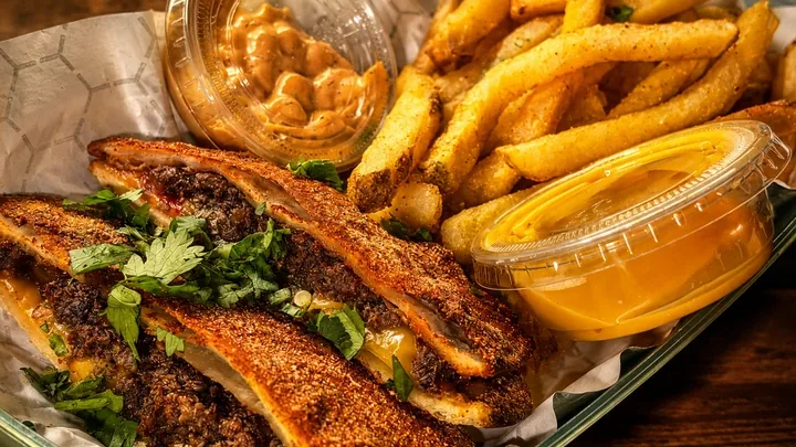 A toasted sandwich cut into strips, filled with dark crumbled filling and melted cheese, served with seasoned fries and two dipping sauces.