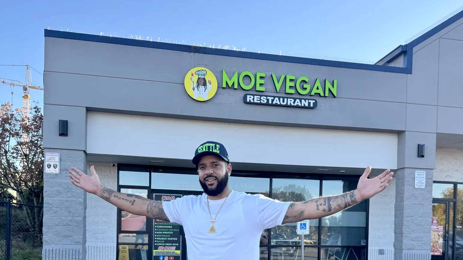 A man in a Seattle cap stands with his arms spread wide in front of the Moe Vegan Restaurant storefront, under its green sign.