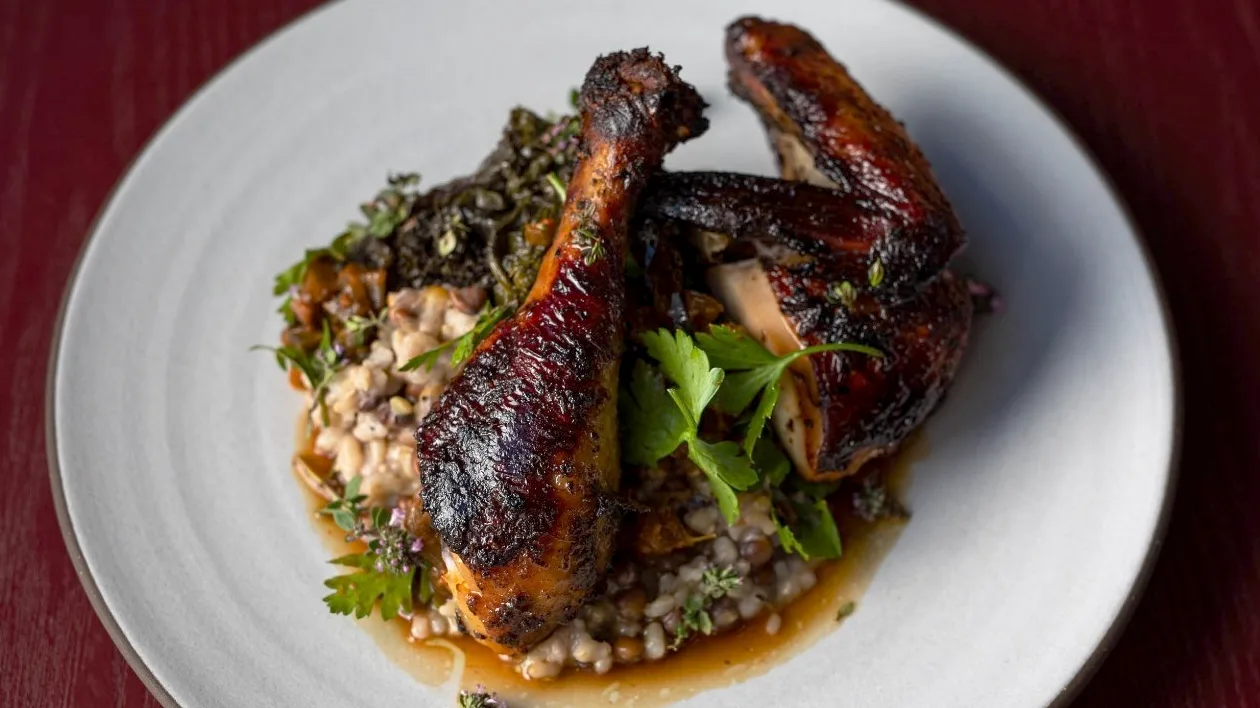 Charred, glazed chicken leg and breast on a bed of grains with braised greens, parsley and thyme flowers in a pool of sauce, on a white plate on a red table.