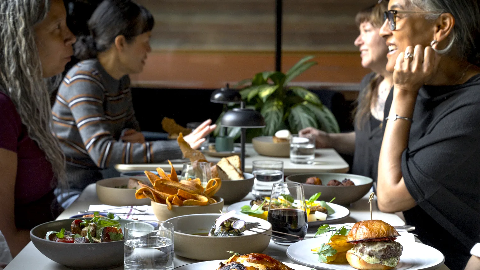 Diners talk and laugh across a long table set with roast chicken, a burger, sweet potato chips, salads and glasses of red wine.